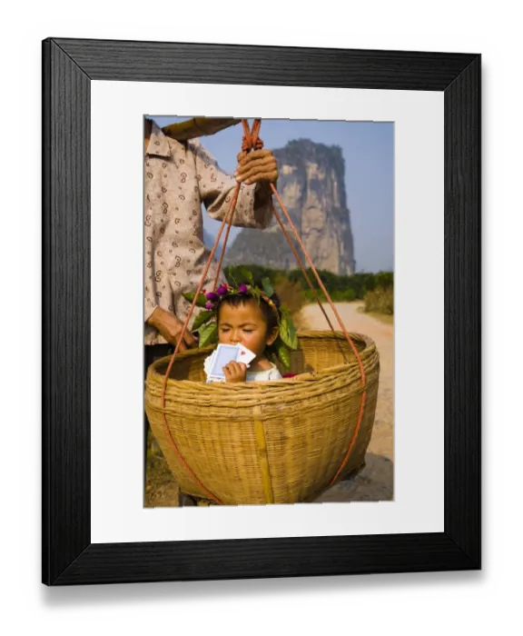 Framed Print : China, Guangxi Zhuang Autonomous Region, Yangshuo County. Young chinese child being carried in an hand woven basket along a rural road running through an agriculture landscape dominated by the Karst peaks of
