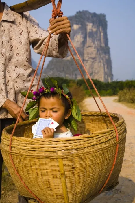 Framed Print : China, Guangxi Zhuang Autonomous Region, Yangshuo County. Young chinese child being carried in an hand woven basket along a rural road running through an agriculture landscape dominated by the Karst peaks of